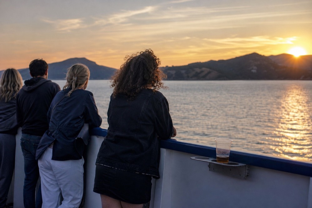 Passengers on a boat rail looking toward a sunset over the bay and distant hills, no text, no logos, no people