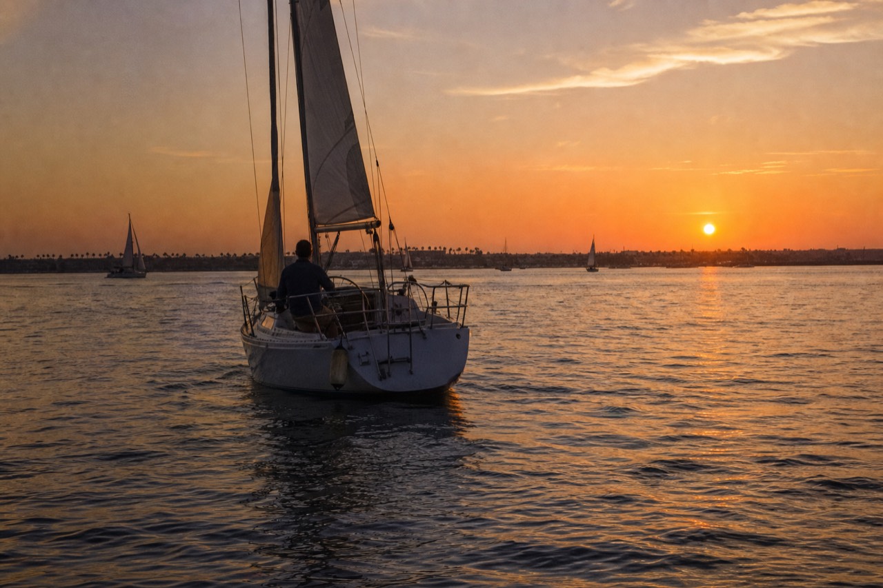 A sailboat crossing San Diego Bay under an orange sunset sky, no text, no logos, no people