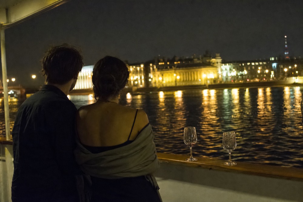 A couple watching the city lights from the open deck of a dinner cruise boat at night, no text, no logos, no people