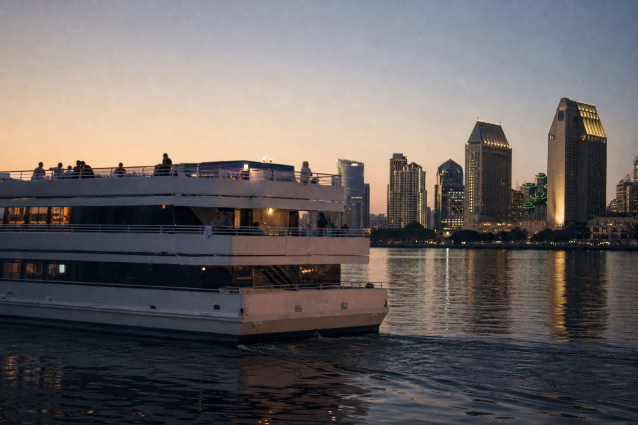 A dinner cruise boat on San Diego Bay at dusk with the downtown skyline behind it, no text, no logos, no people