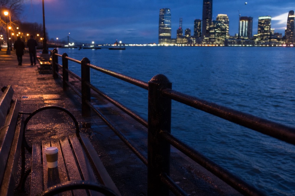 Empty waterfront path along the bay at twilight with city lights beginning to glow, no text, no logos, no people