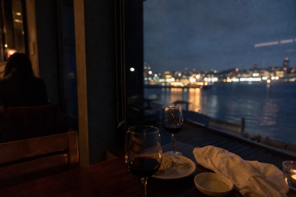 A dimly lit dining table with wine glasses beside a window reflecting harbor lights, no text, no logos, no people