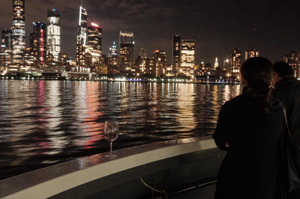 A city skyline glowing across dark water as seen from a boat deck at night, no text, no logos, no people