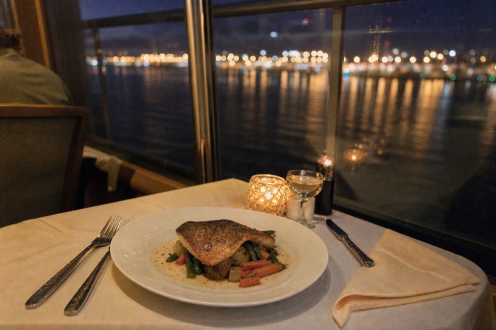 A plated dinner course on a cruise ship table with the bay lights in the background, no text, no logos, no people