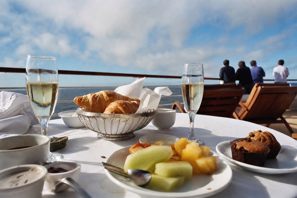 A brunch buffet spread with pastries and fruit beside glasses of sparkling cider on a sunlit cruise deck, no text, no logos, no people