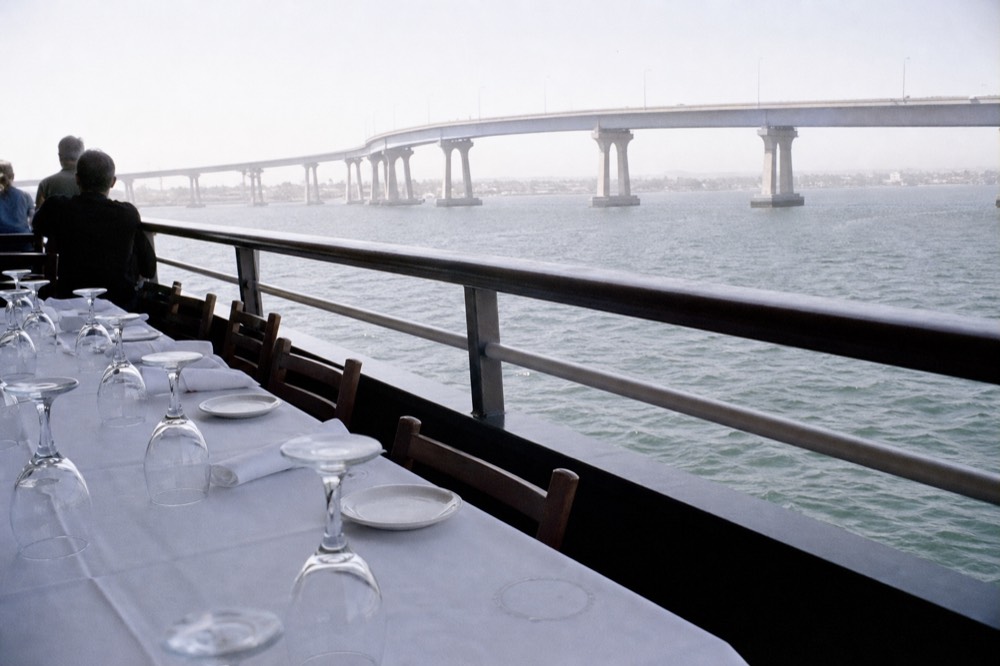 A group of place settings at a long table on a cruise deck with the Coronado Bridge in the distance, no text, no logos, no people