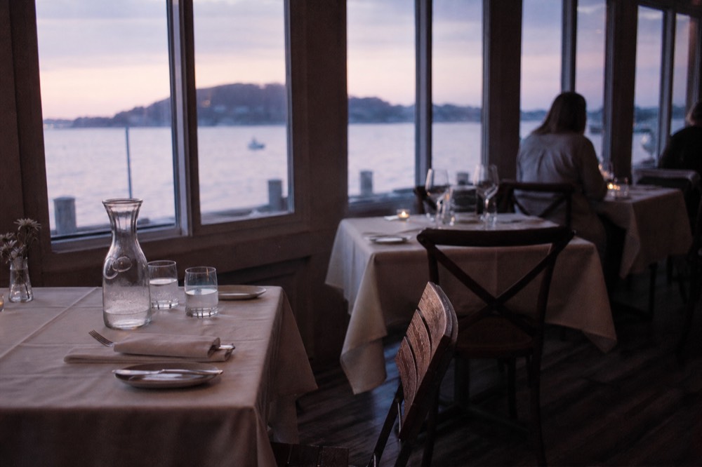 Indoor dining salon with set tables and wide windows overlooking the bay at dusk, no text, no logos, no people