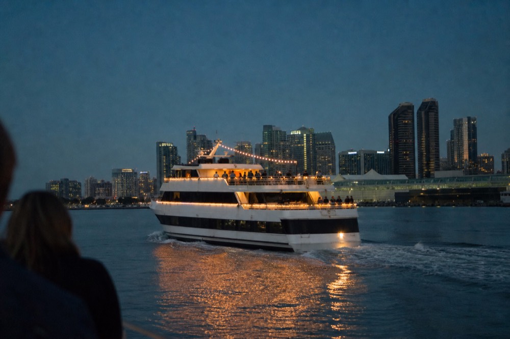 Dinner cruise vessel lit at dusk moving across San Diego Bay with the city skyline behind, no text, no logos, no people