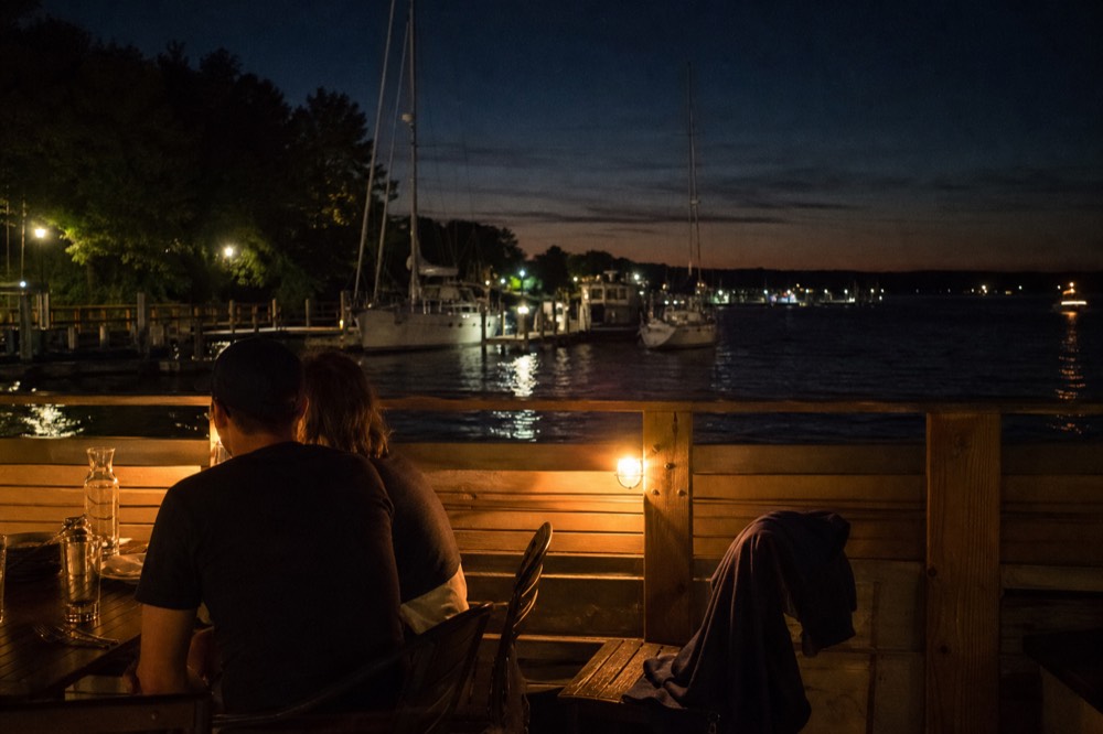 Evening waterfront scene with diners on an outdoor patio near sailboats, no text, no logos, no people