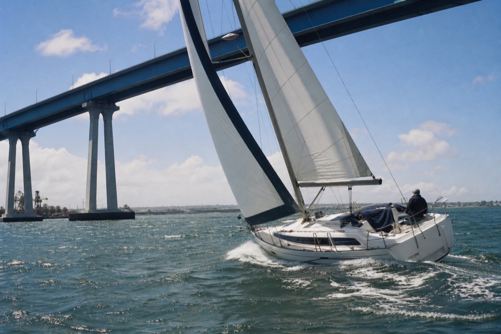 A small sailboat heeling on San Diego Bay with the Coronado Bridge in the background, no text, no logos, no people