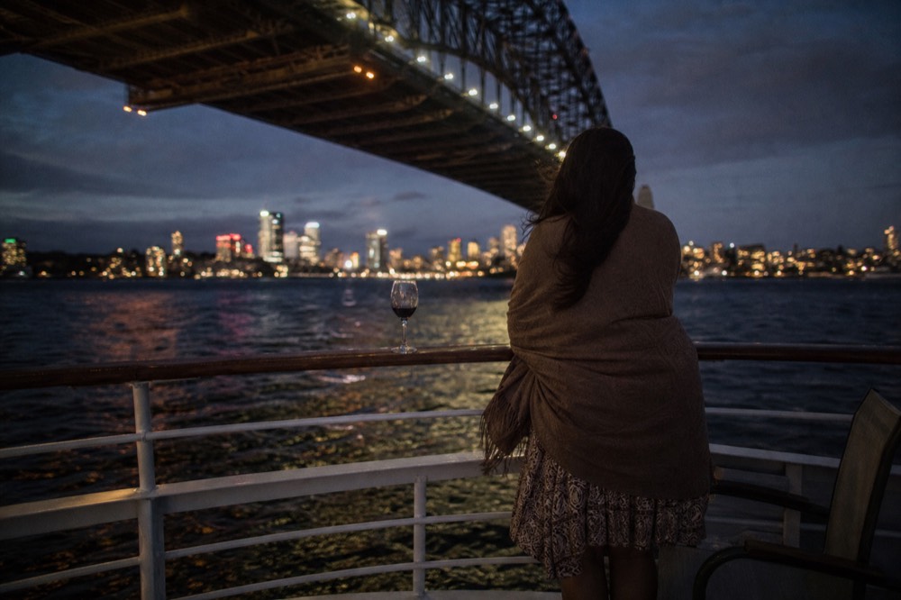 A woman in a shawl and dress standing at a ship railing watching city lights across dark water, no text, no logos, no people