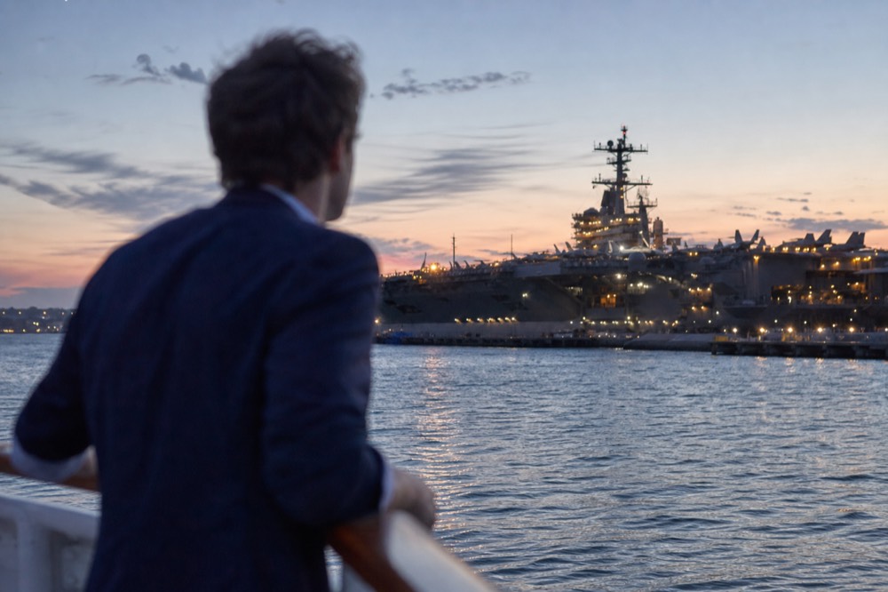 A man in a blazer and collared shirt leaning on a ship rail at dusk, no text, no logos, no people