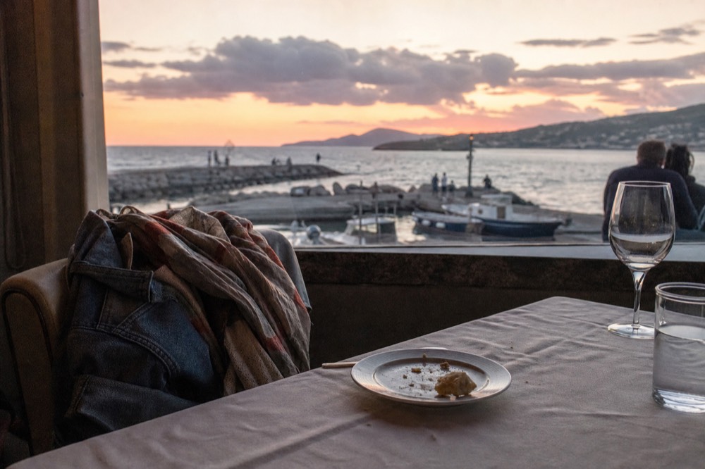 A folded jacket and scarf resting on a chair at a set dinner table beside a window showing dusk over water, no text, no logos, no people