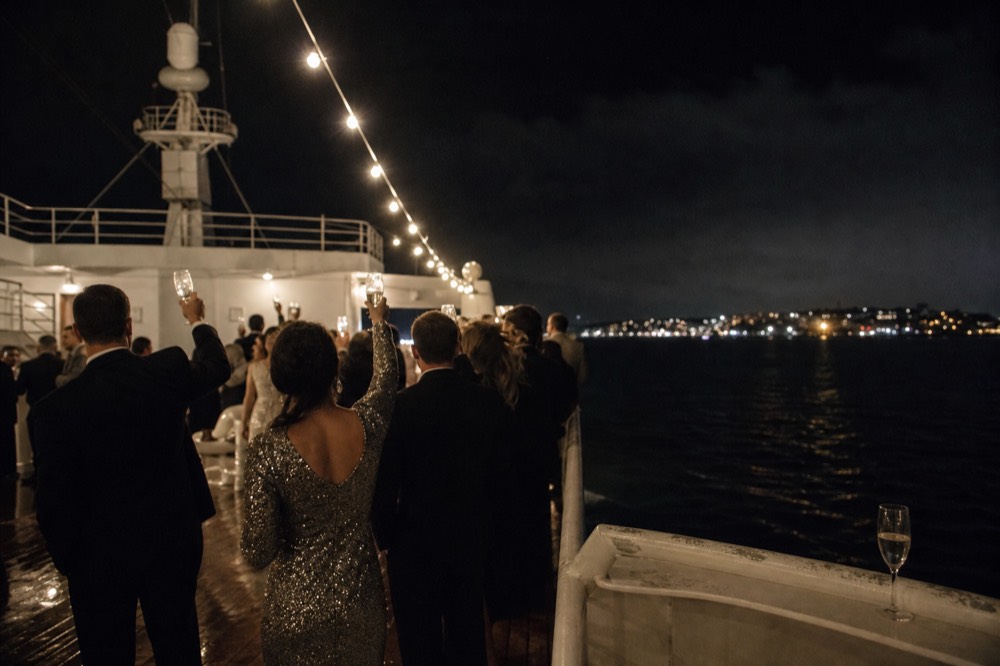 Sparkling dresses and dark suits on passengers toasting under string lights on an outdoor ship deck at night, no text, no logos, no people