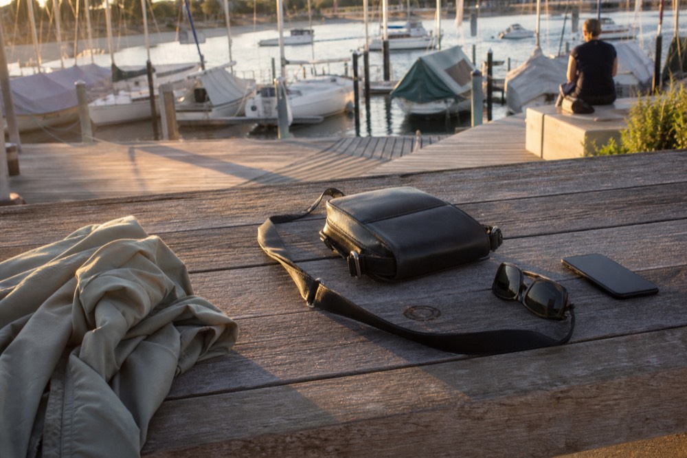 A small crossbody bag, sunglasses, a light jacket and a phone arranged on a bench by a marina at golden hour, no text, no logos, no people